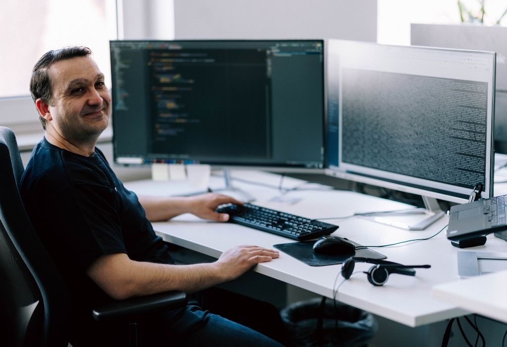 Freundlich lächelnder Mann sitzt an einem modernen Arbeitsplatz vor mehreren Monitoren im Büro und steht sinnbildlich für professionelle Magento-Beratung und technische E-Commerce-Expertise